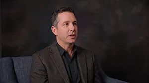 Example video shoot frame: a man in a gray blazer seated in front of a dark canvas backdrop